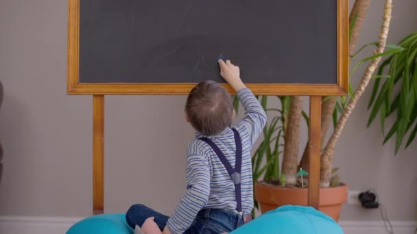 Back view portrait of autistic cheerful boy writing Mama at blackboard with chalk laughing looking away. Wide shot happy motivated Caucasian child with mental disorder studying in comfortable school. — стоковое видео