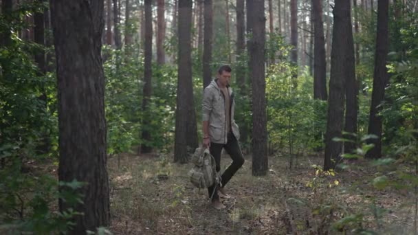 Wide shot woodland landscape with Caucasian man walking with backpack leaving behind the tree. Confident male traveler hiking outdoors in spring summer forest. Backpacking and tourism. Slow motion. — Stockvideo