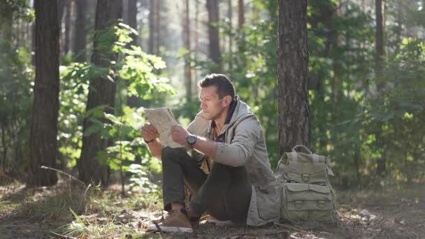 Wide shot portrait of focused Caucasian man sitting in forest examining paper map. Confident concentrated male traveler choosing direction resting in sunshine on spring summer day. Adventure concept. — стоковое видео