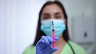 Syringe with coronavirus vaccine in hand of blurred female doctor in Covid-19 face mask. Close-up pandemic illness treatment in injection pen with serious professional woman at background.