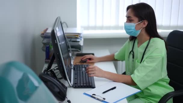 集中的专业医生上网在电脑上坐在室内餐桌边。在医院工作的身穿制服和科维德面罩的年轻白人妇女的侧视图画像. — 图库视频影像