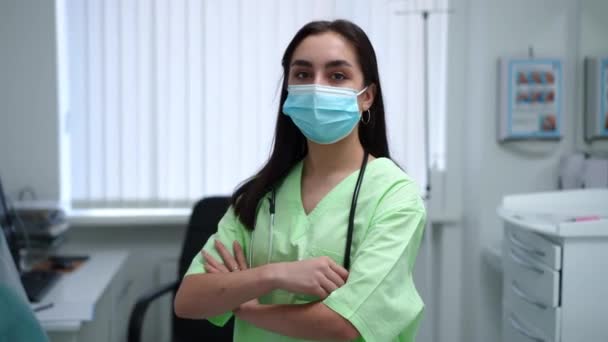 Confident woman in coronavirus face mask and green medical uniform crossing hands looking at camera standing indoors. Portrait of professional Caucasian female doctor posing in clinic. — Video
