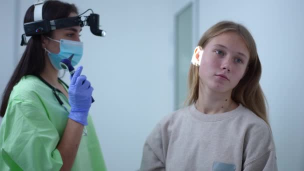 Teenage Caucasian girl sitting in hospital as otolaryngologist with otoscope entering checking ears. Portrait of teen patient and professional female doctor indoors. Medical examination concept. — стоковое видео