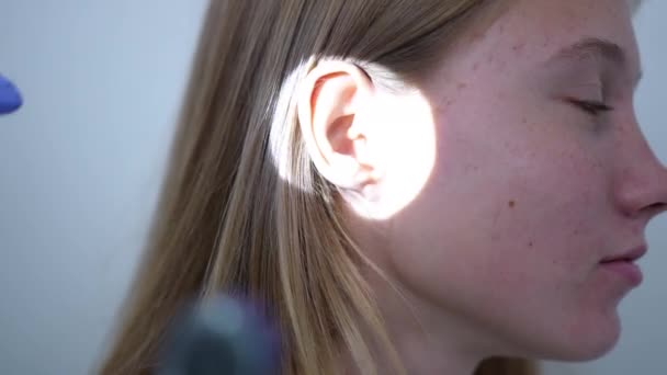 Close-up orelha vista lateral de paciente adolescente do sexo feminino com foco rack ao otoscópio na mão do médico. Otorrinolaringologista irreconhecível que verifica o canal auditivo e membrana timpânica no hospital dentro de casa. — Vídeo de Stock