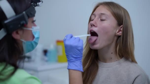 Portrait of teenage Caucasian girl opening mouth for medical examination as blurred doctor checking throat and tongue. Cute teenager undergoing checkup of otolaryngologist. — Stockvideo