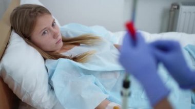 Sad ill teenage girl lying in hospital ward with nurse hands adjusting dropper. High angle view portrait of devastated depressed Caucasian teenager undergoing treatment in medical clinic.