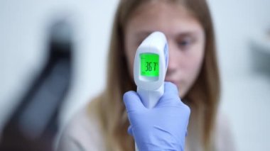 Close-up contactless thermometer showing 36.7 degrees with blurred teenage Caucasian girl at background. Unrecognizable doctor nurse measuring patient temperature in hospital indoors.