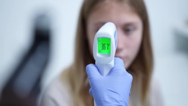 Närbild kontaktlös termometer visar 36,7 grader med suddig tonåring kaukasiska flicka i bakgrunden. Oigenkännlig läkare sjuksköterska mäter patientens temperatur på sjukhus inomhus. — Stockvideo