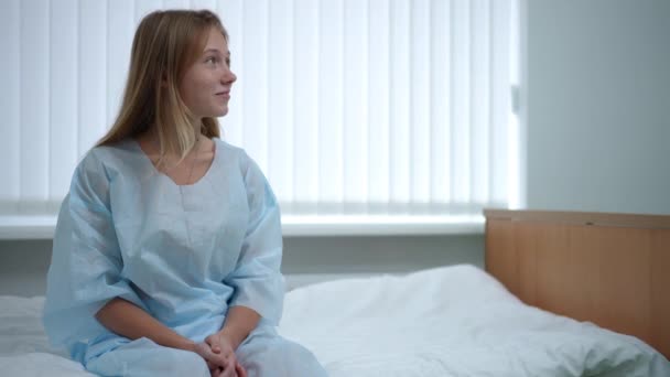 Teenage girl sitting on bed in hospital ward as doctor entering talking smiling. Portrait of positive Caucasian patient with professional physician in medical clinic indoors. Recovery and overcome. — Wideo stockowe