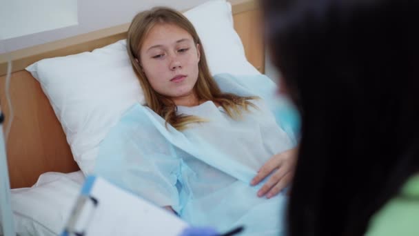 Portret nastolatki leżącej w łóżku z kroplomierzem, mówiącej narzekającym na ból. Niewyraźny lekarz spisujący objawy siedzące na boku. Smutny pacjent rasy kaukaskiej na oddziale szpitalnym z pielęgniarką. — Wideo stockowe
