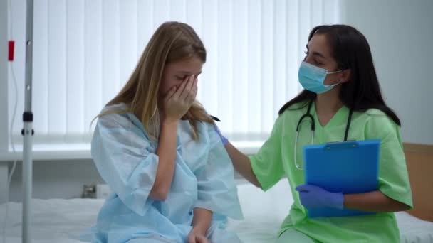 Nastolatka płacząca na łóżku z lekarzem dotykającym ramienia popierającym pacjenta. Portret zdewastowanej, przygnębionej nastolatki rasy kaukaskiej otrzymującej wiadomości o śmiertelnej chorobie od kobiety w klinice. — Wideo stockowe