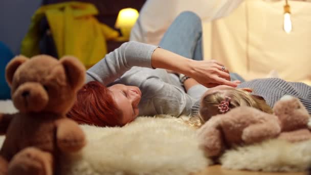 Smiling Caucasian mother caressing face of little daughter in slow motion lying at tent in living room. Happy woman stroking charming girl enjoying tranquility at home indoors on weekend. — стоковое видео