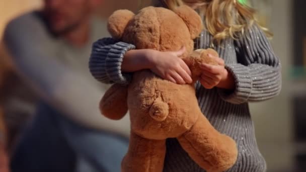 Close-up teddy bear in hands of unrecognizable little girl with blurred man at background. Caucasian child playing with toy in slow motion resting at home with father. Childhood and family. — Wideo stockowe