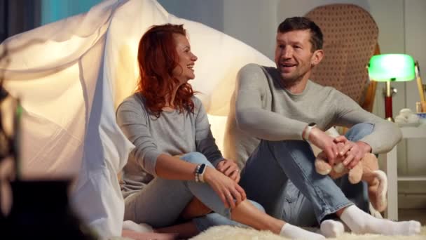 Ampla tiro feliz casal adulto abraçando sentado na tenda na sala de estar falando em câmera lenta sorrindo. Homem e mulher caucasianos amorosos descansando dentro de casa no fim de semana. Romance e amor. Movimento lento. — Vídeo de Stock