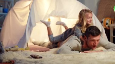 Wide shot joyful man imitating horse riding with cute little girl on back. Portrait of happy Caucasian smiling father playing with daughter in slow motion at tent in living room. Family leisure joy.
