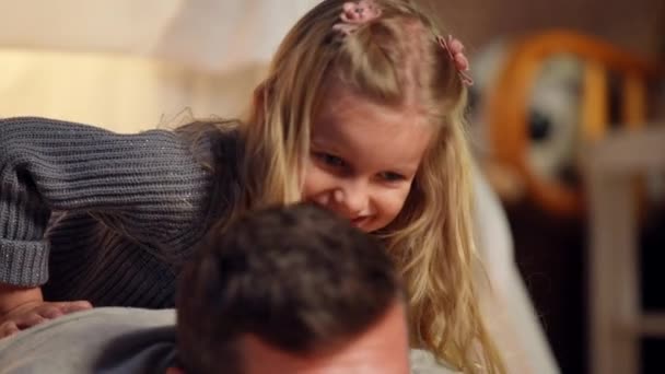 Close-up charming excited daughter looking away lying on back of father imitating riding in slow motion. Live camera moves down to Caucasian man playing with pretty girl on weekend at home. — стокове відео