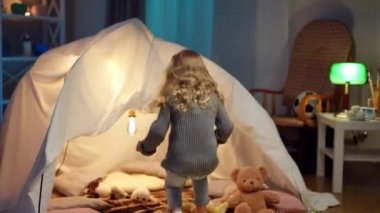 Tracking shot of cheerful little girl running to tent in living room sitting inside. Happy positive Caucasian child having fun at night at home indoors. Live camera follows kid.