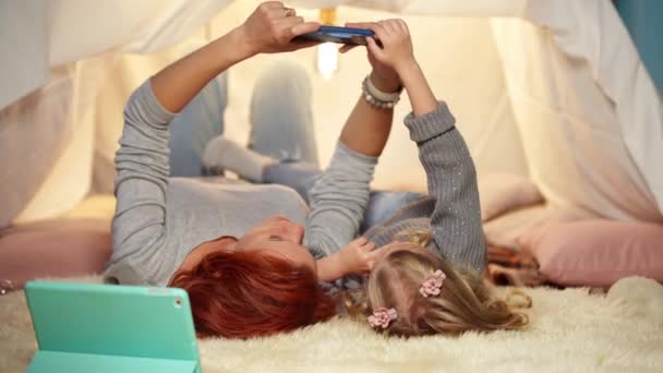 Relaxed mother and little daughter taking selfie on smartphone lying in cozy tent in living room. Happy Caucasian woman and girl resting at home indoors talking in slow motion. — стоковое видео
