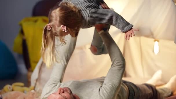 Happy excited girl looking around imitating flying as man raising kid in slow motion. Portrait of cheerful Caucasian daughter enjoying weekend leisure with father at tent in living room. — Stok Video