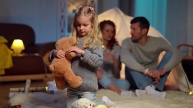Wide shot portrait of serious pretty girl playing with teddy bear as blurred father and mother talking at background. Confident Caucasian daughter resting with man and woman at home in living room.