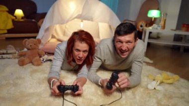 Front view laughing joyful couple gaming with game controllers looking at camera. Positive happy carefree Caucasian man and woman having fun enjoying hobby at tent in living room at home.