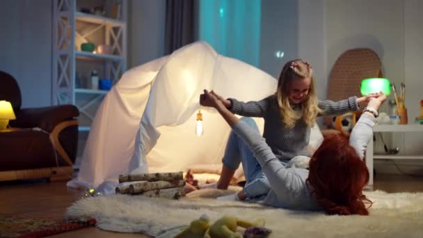 Wide shot cheerful little daughter jumping on mother lying at tent in living room. Joyful happy Caucasian girl having fun enjoying leisure with woman indoors at home. Hyperactivity and parenting. — Vídeo de stock