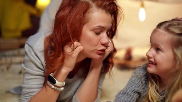 Front view portrait of beautiful mother kissing nose of cute daughter laughing. Happy Caucasian woman enjoying leisure with pretty carefree girl at tent in living room at home indoors. — Video Stock