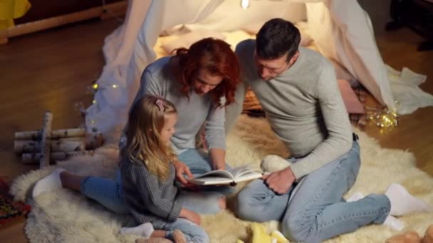 Wysoki kąt widzenia kaukaski ojciec matka i córka rozmawiają książki w namiocie w salonie. Szeroki strzał zrelaksowany mężczyzna kobieta i dziewczynka korzystających odpoczynku przed snem w domu. — Wideo stockowe