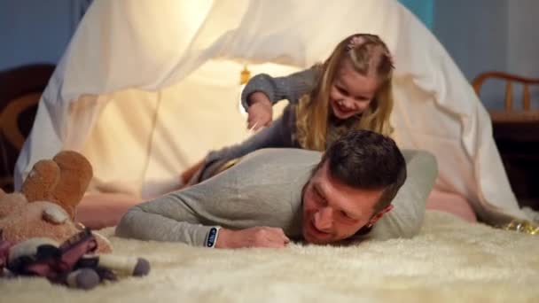 Front view cheerful father laughing as daughter hugging parent lying on back in tent in living room. Portrait of happy Caucasian man having fun enjoying leisure with little cute girl indoors at home. — Vídeo de stock