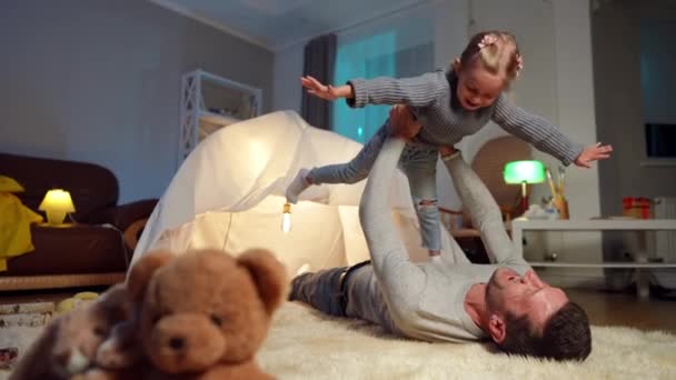 Happy daughter imitating flying in father hands and hugging parent smiling lying at tent in living room. Wide shot portrait of joyful Caucasian girl enjoying leisure with man at home on Fathers Day. — Vídeo de stock