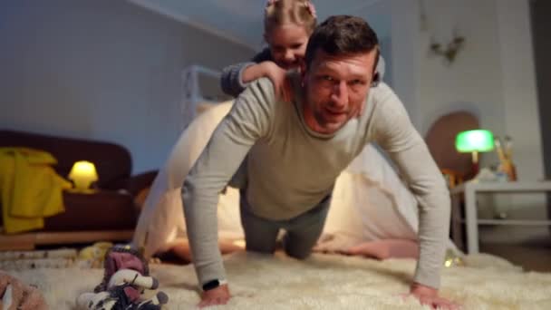 Athletic Caucasian father doing pushups looking at camera with cute little daughter on back. Portrait of happy strong man training at home in living room with curios girl. Lifestyle and parenting. — Wideo stockowe