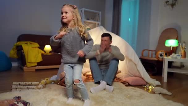 Joyful cute girl dancing in living room at home with blurred man clapping laughing sitting at tent at background. Portrait of cheerful daughter having fun enjoying weekend leisure with father. — Videoclip de stoc