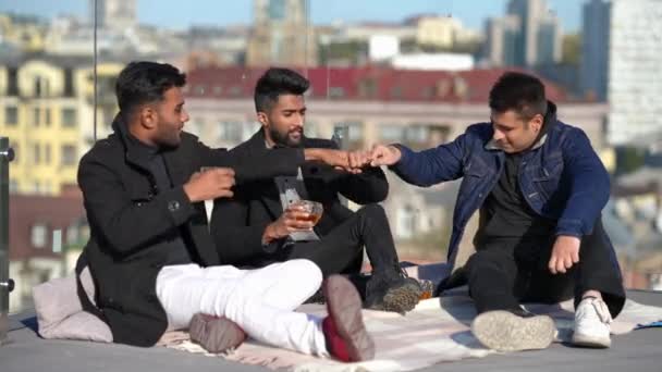Wide shot relaxed Middle Eastern groom celebrating bachelor party with friends on rooftop. Happy handsome young men bumping fists clinking whiskey glasses talking smiling with urban city at background — Video Stock