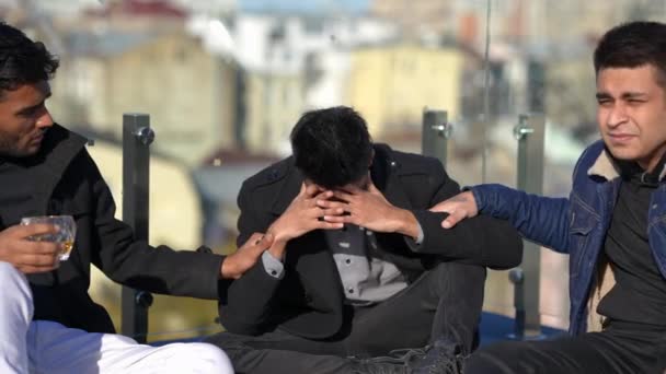 Young devastated Middle Eastern man looking at camera sitting with friends on sunny rooftop outdoors. Depressed frustrated handsome guy posing as mates endorsing stressed friend. Friendship concept. — Video