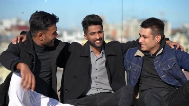 Tres felices hombres del Medio Oriente saludando mirando a la cámara sonriendo sentados en una cerca de cristal en la azotea. Retrato de amigos relajados guapos confiados posando al aire libre en la ciudad urbana. — Vídeo de stock