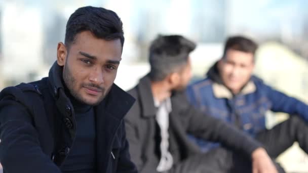 Portrait of handsome young man looking at camera smiling as blurred friends talking at background. Confident Middle Eastern guy posing outdoors partying with mates on rooftop in urban city. — Stockvideo