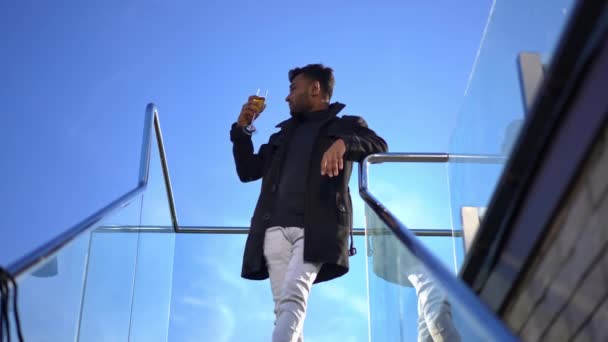 Wealthy confident Middle Eastern young man drinking whiskey standing at stairs on rooftop looking away. Portrait of rich successful businessman enjoying sunny day in penthouse outdoors. — Video Stock