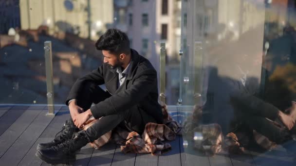 Wide shot anxious troubled Middle Eastern man sitting at glass fence on rooftop looking away. Portrait of worried stressed depressed handsome guy thinking resting outdoors on sunny day. — Stockvideo