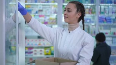 Yan görüş pozitif eczane çalışanı ilaçlarını raflara koyuyor gülümsüyor ve gidiyor. Arka planda uyuşturucuyu seçen bulanık müşteriler. İçerideki eczanede çalışan beyaz bir kadın..