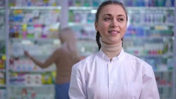 Jeune femme souriante belle debout dans la pharmacie regardant autour et regardant la caméra. Portrait de pharmacienne caucasienne confiante posant à l'intérieur avec des clients flous en arrière-plan. — Video