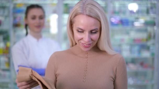 Portret van tevreden vrouwelijke cliënt poseren met support kousen in de apotheek met wazig drogist op de achtergrond. Front view gelukkig glimlachen Kaukasische vrouw kijken naar camera reclame panty 's. — Stockvideo