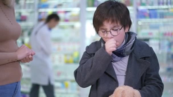 Unwell Caucasian boy coughing standing in pharmacy with unrecognizable woman caressing hair. Portrait of ill son with mother in drugstore buying remedies. Cold and flu treatment concept. — ストック動画