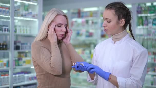 Sad ill woman complaining headache to pharmacist passing pills in blister. Portrait of Caucasian customer and druggist passing medication standing indoors in drugstore talking. Migraine treatment. — стоковое видео