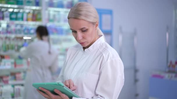 Portrait of intelligent focused woman checking online list of medications comparing stock on shelves in drugstore. Blurred colleague standing at background. Concentrated Caucasian druggist in pharmacy — стоковое видео