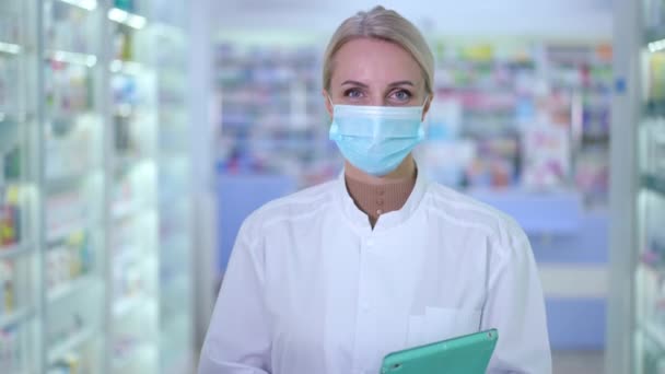 Портрет улыбающейся женщины в маске Ковида, смотрящей на камеру, стоящую с планшетом в аптеке. Уверенный в себе красивый кавказский аптекарь, позирующий в помещении аптеки в связи с пандемией коронавируса. — стоковое видео