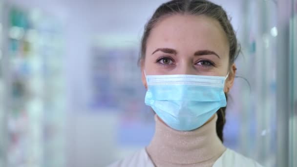 Close-up face of smiling woman in coronavirus face mask on the right in pharmacy indoors. Portrait of confident attractive Caucasian druggist posing in drugstore on Covid-19 pandemic outbreak. — Video