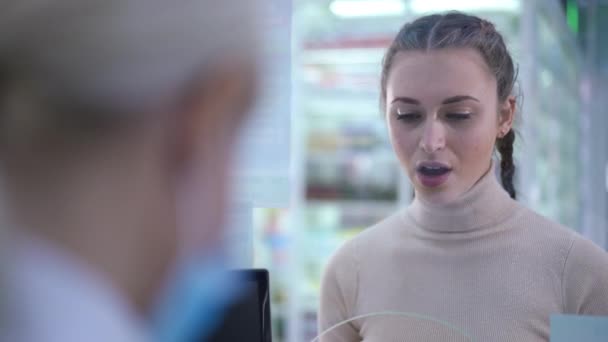 Portret van een jonge slanke, mooie vrouw die drugsverslaafd advies geeft aan de kassa in de apotheek. Geconcentreerde blanke cliënt in slow motion in drogisterij in gesprek met apotheker. — Stockvideo