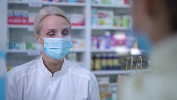 Positive expert female pharmacist smiling talking with client at cashdesk in drugstore. Portrait of beautiful Caucasian woman in Covid face mask consulting customer in pharmacy indoors. — Vídeos de Stock