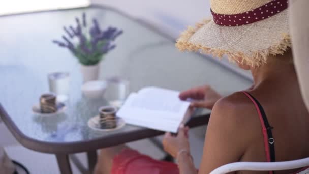 Shooting over shoulder of confident Caucasian woman reading turning pages in slow motion sitting at table with coffee cups. Intelligent slim elegant lady enjoying hobby on summer morning on terrace. — Vídeo de Stock