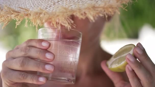 Close-up front view unrecognizable slim woman in straw hat drinking refreshing ice water holding lemon half in hand. Confident Caucasian lady enjoying summer breakfast on terrace outdoors. — стоковое видео